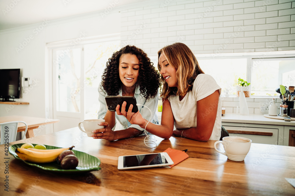 Two caucasian friends sitting in kitchen catching up watching videos on cellular device Two caucasian friends sitting in kitchen catching up watching videos on cellular device