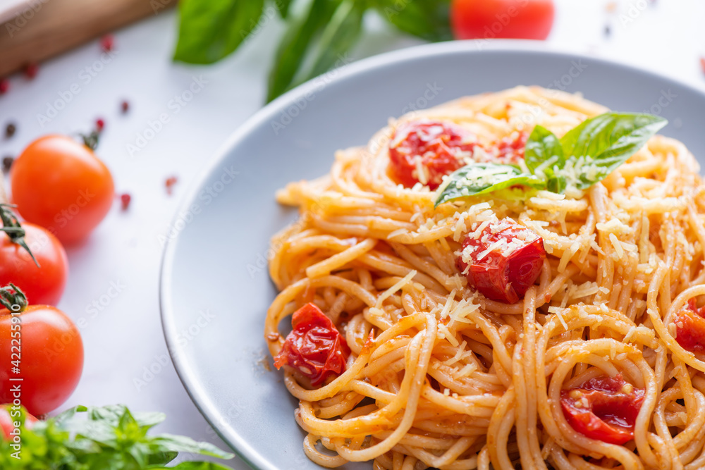Tasty appetizing classic italian spaghetti pasta with tomato sauce, cheese parmesan and basil on plate and ingredients for cooking pasta on white marble table.