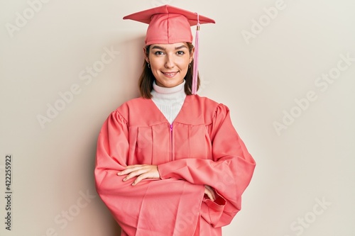 Young caucasian woman wearing graduation cap and ceremony robe happy face smiling with crossed arms looking at the camera. positive person.