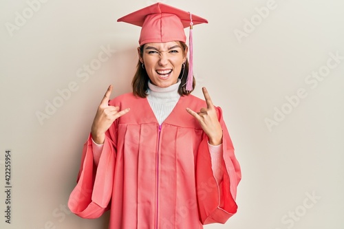 Young caucasian woman wearing graduation cap and ceremony robe shouting with crazy expression doing rock symbol with hands up. music star. heavy concept.