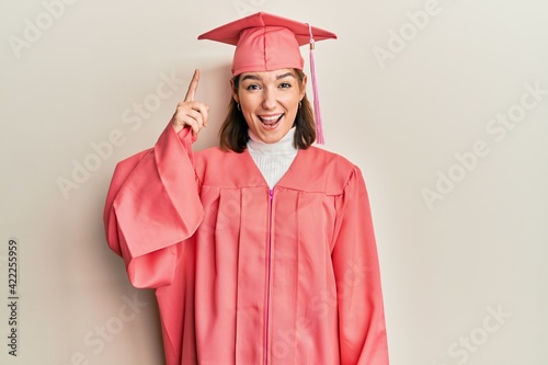 Young caucasian woman wearing graduation cap and ceremony robe pointing finger up with successful idea. exited and happy. number one.