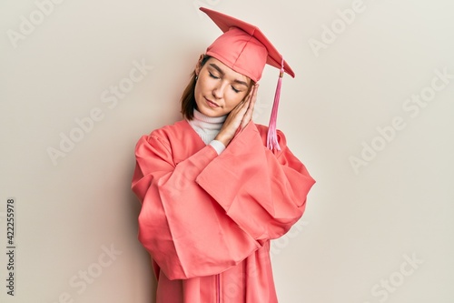 Young caucasian woman wearing graduation cap and ceremony robe sleeping tired dreaming and posing with hands together while smiling with closed eyes.