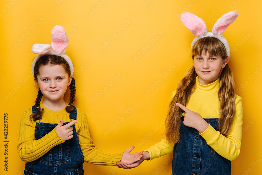 Portrait of happy two cute little girls in pink bunny fluffy ears, points for finger aside, demonstrates something over blank space, isolated over yellow studio background. Easter holiday concept Portrait of happy two cute little girls in pink bunny fluffy ears, points for finger aside, demonstrates something over blank space, isolated over yellow studio background. Easter holiday concept