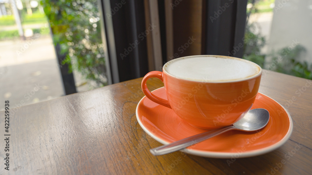 Orange cup of hot cappuccino coffee on wooden table within cafe near glass window, modern lifestyle concept Orange cup of hot cappuccino coffee on wooden table within cafe near glass window, modern lifestyle concept