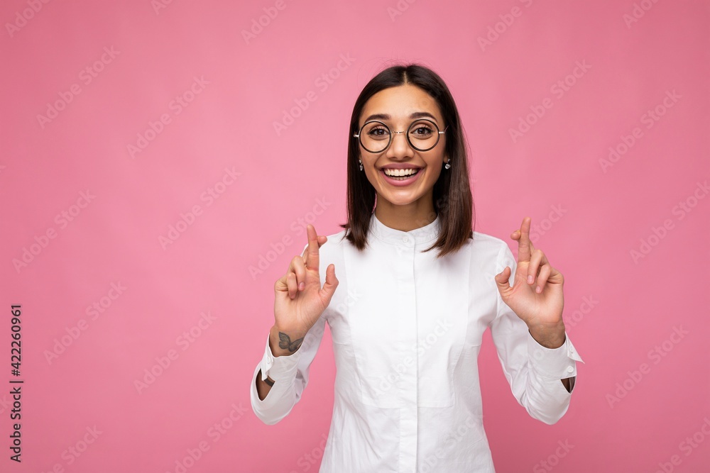 Portrait of young positive happy attractive brunette woman with sincere emotions wearing casual white shirt and optical glasses isolated over pink background with copy space and holding fingers Portrait of young positive happy attractive brunette woman with sincere emotions wearing casual white shirt and optical glasses isolated over pink background with copy space and holding fingers