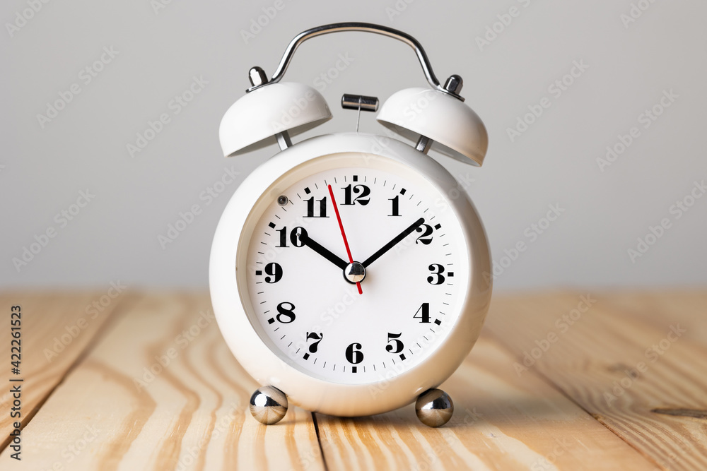 Small white alarm clock, black numbers, set the time for 10.10 o'clock, placed on a wood table. Small white alarm clock, black numbers, set the time for 10.10 o'clock, placed on a wood table.