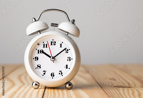 Small white alarm clock, black numbers, set the time for 10.10 o'clock, placed on a wood table.