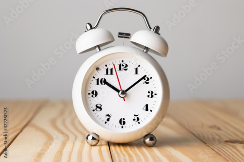 Small white alarm clock, black numbers, set the time for 10.10 o'clock, placed on a wood table.