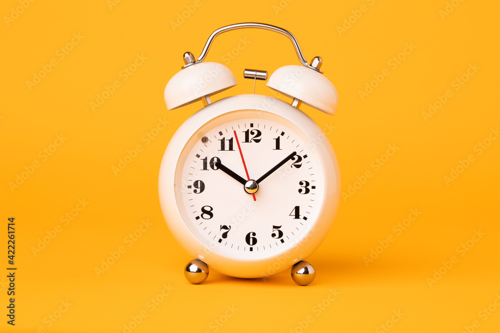 Small white alarm clock, black numbers, set the time placed on a table. Clock on isolated yellow background. Small white alarm clock, black numbers, set the time placed on a table. Clock on isolated yellow background.