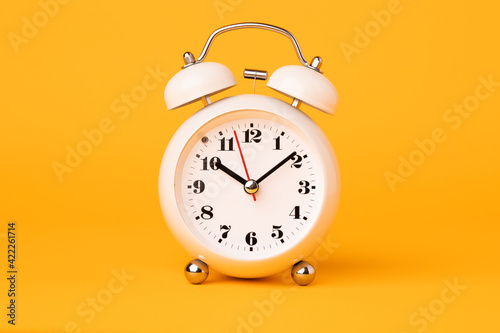 Small white alarm clock, black numbers, set the time placed on a table. Clock on isolated yellow background.