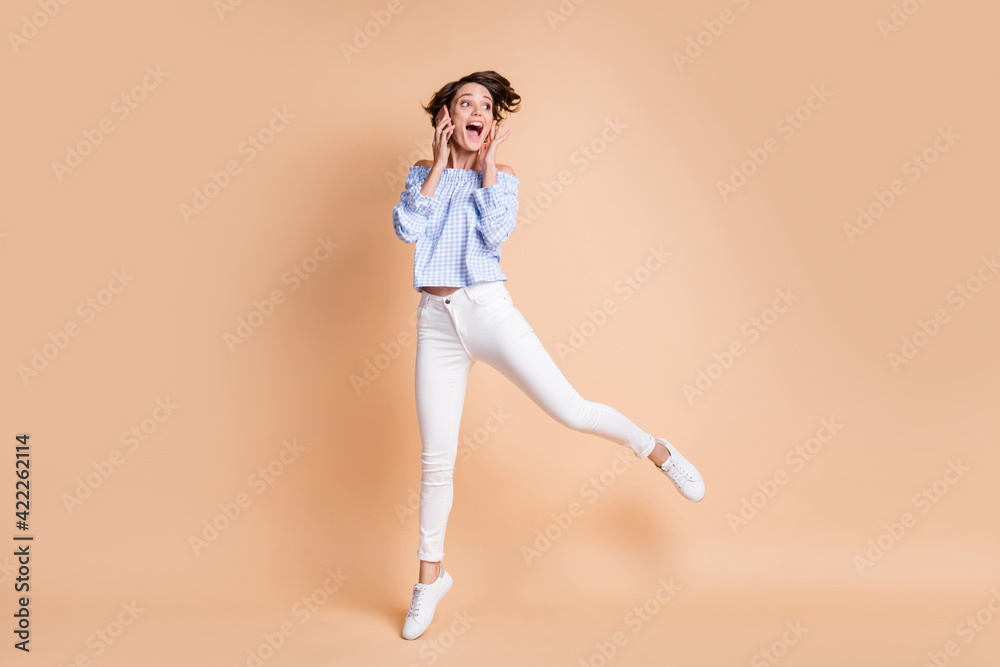 Full size photo f young excited girl happy amazed surprised talk cellphone jump look empty space isolated over beige color background Full size photo f young excited girl happy amazed surprised talk cellphone jump look empty space isolated over beige color background