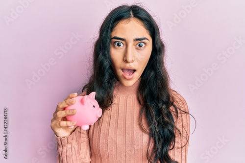 Hispanic teenager girl with dental braces holding piggy bank scared and amazed with open mouth for surprise, disbelief face © Krakenimages.com
