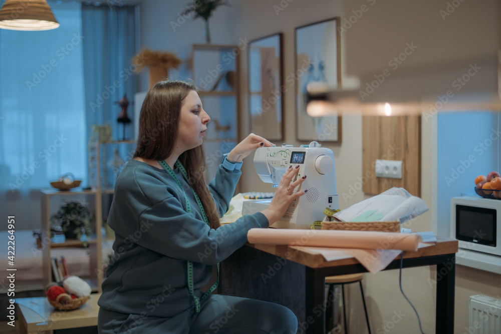 Young woman sews on a sewing machine in the living room at home