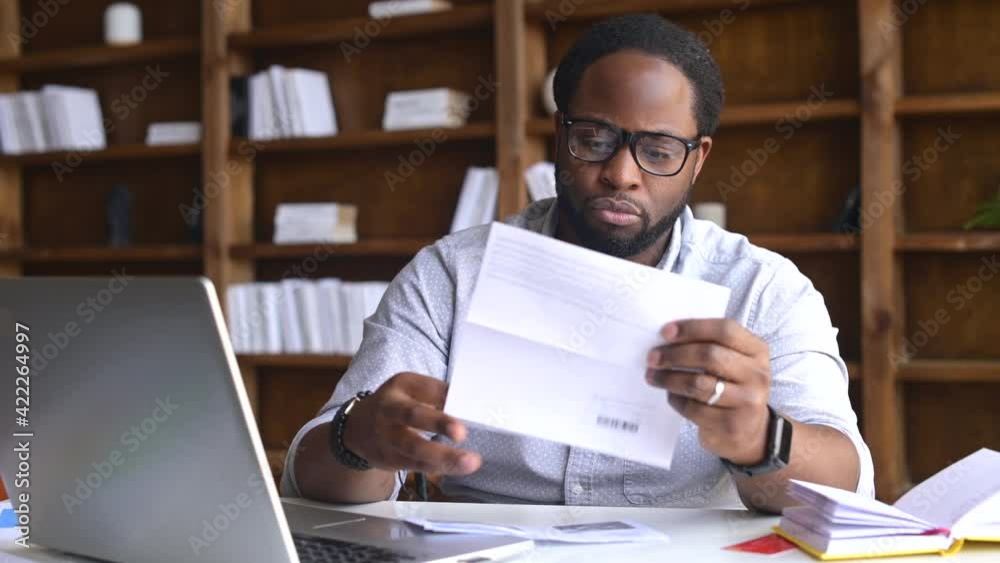 Serious African-American businessman looking through mailing correspondence, concentrated man opens envelope and take letter out, reading a document, received an important notification by mail