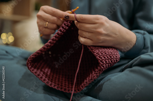 Young woman crochets a basket on the sofa in the living room. Close-up of hands.