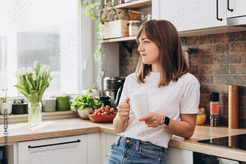 Close up woman in white t-shirt drinking coffee or tea in kitchen at home. Early breakfast in the morning, rest time