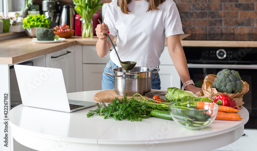 Close up woman in white t-shirt cooking soup with fresh vegetables in kitchen at home. Menu  recipe book banner. Girl reads the recipe in laptop. Caucasian model using internet in loft apartment.