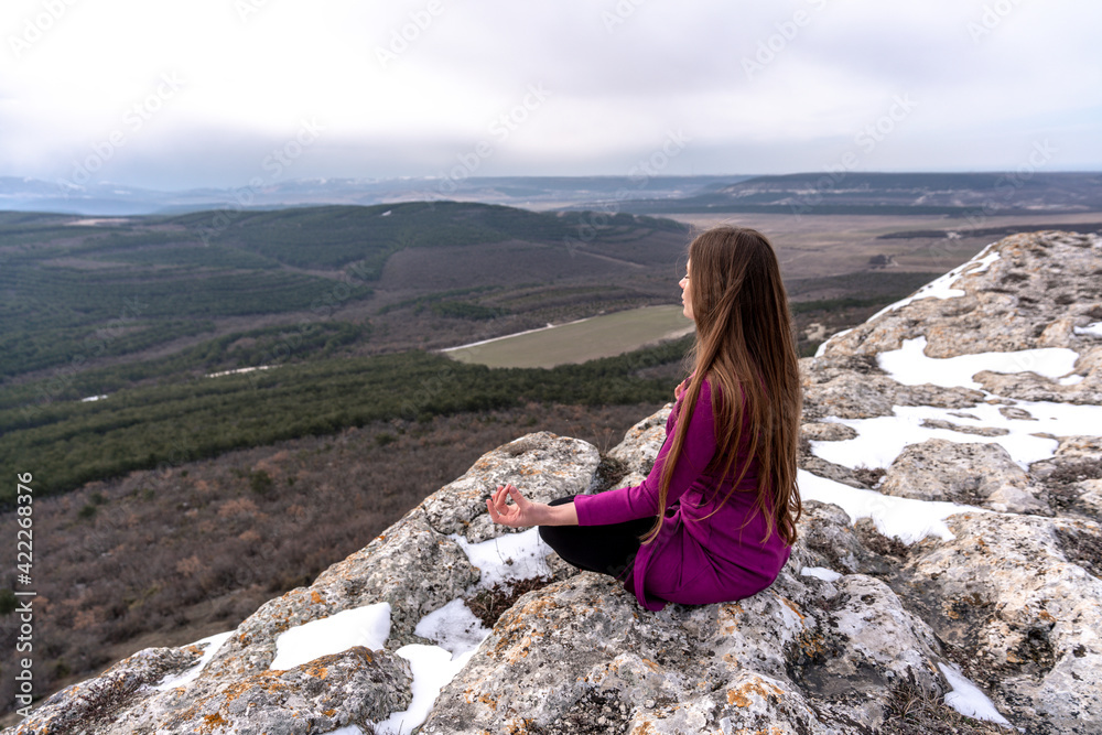 Naklejka premium A beautiful woman sits in a lotus position on a high place with an amazing view of the mountains and the gorge practicing yoga meditation Kundalini energy thinking intuition prana. Loneliness harmony
