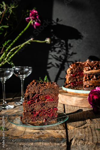 Piece of homemade chocolate and cherry cake  whole cake  on wooden stand  glasses and flowers on dark background.