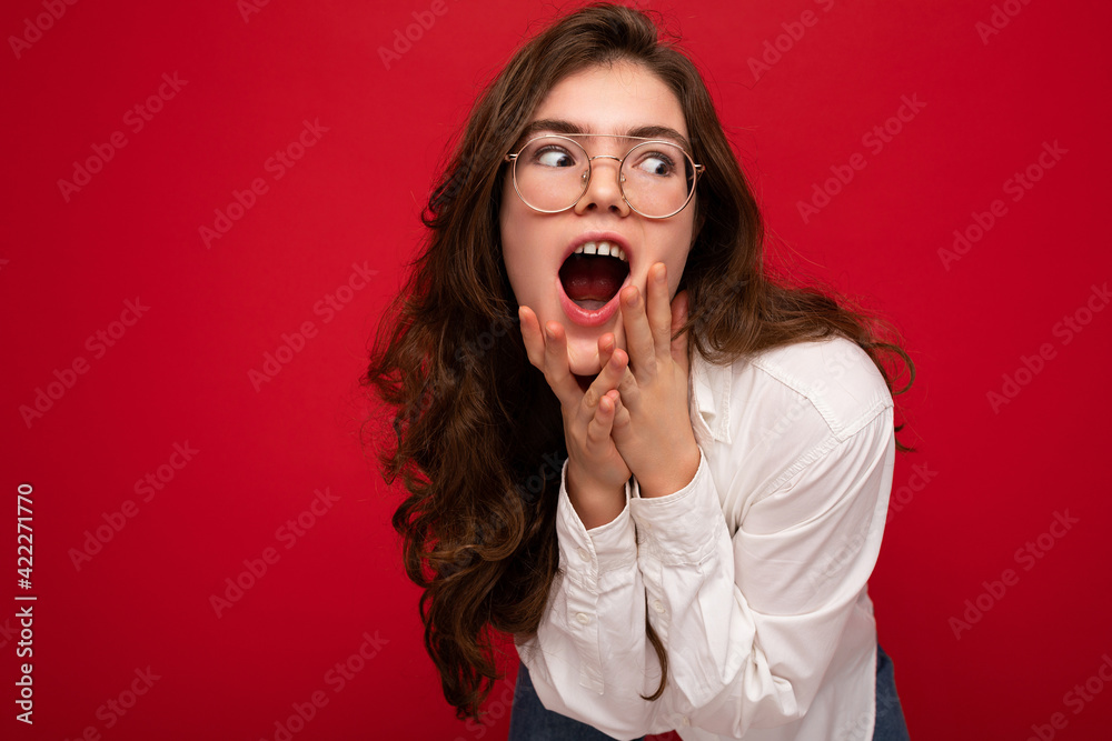 Photo shot of beautiful shocked young brunette curly woman isolated over red background wearing casual white shirt and stylish optical glasses looking to the side with open mouth Photo shot of beautiful shocked young brunette curly woman isolated over red background wearing casual white shirt and stylish optical glasses looking to the side with open mouth