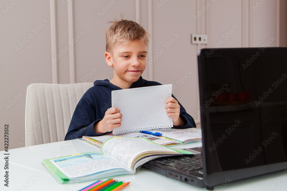 Boy shows notebook with answer to teacher on laptop screen. Smart Little Boy Is doing His Homework in Living Room. COVID-19 pandemic forces children online learning Boy shows notebook with answer to teacher on laptop screen. Smart Little Boy Is doing His Homework in Living Room. COVID-19 pandemic forces children online learning