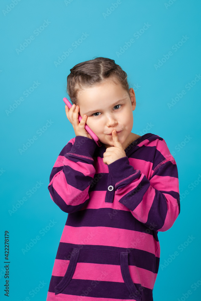 close-up portrait of a mysterious girl with pigtails making a phone call and holding a finger to her mouth, a secret or a request for silence, isolated on a blue background. close-up portrait of a mysterious girl with pigtails making a phone call and holding a finger to her mouth, a secret or a request for silence, isolated on a blue background.