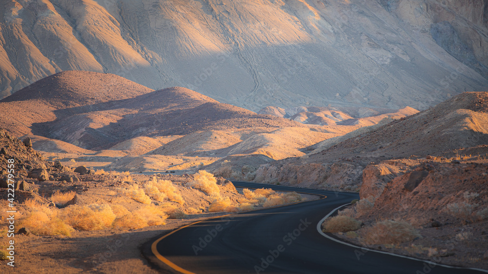 Dramatic golden light on an empty winding desert road through the rugged terrain of the badlands landscape in Death Valley Park National Park, USA.