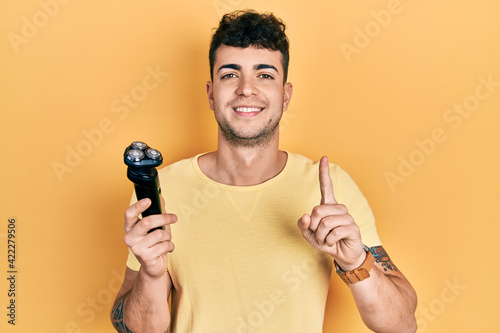 Young hispanic man holding electric razor smiling with an idea or question pointing finger with happy face, number one