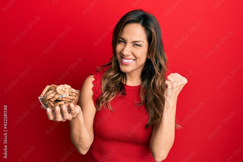 Young latin woman holding bowl with cookies screaming proud, celebrating victory and success very excited with raised arm Young latin woman holding bowl with cookies screaming proud, celebrating victory and success very excited with raised arm