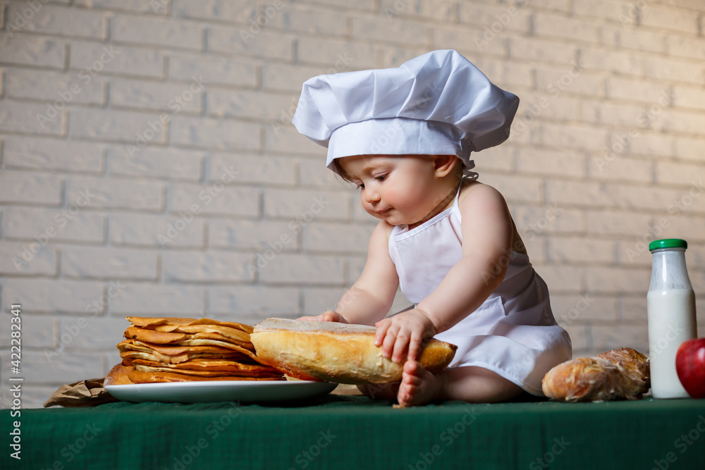 Little boy dressed as a cook in the kitchen. Handsome child dressed in an apron eating bread Little boy dressed as a cook in the kitchen. Handsome child dressed in an apron eating bread