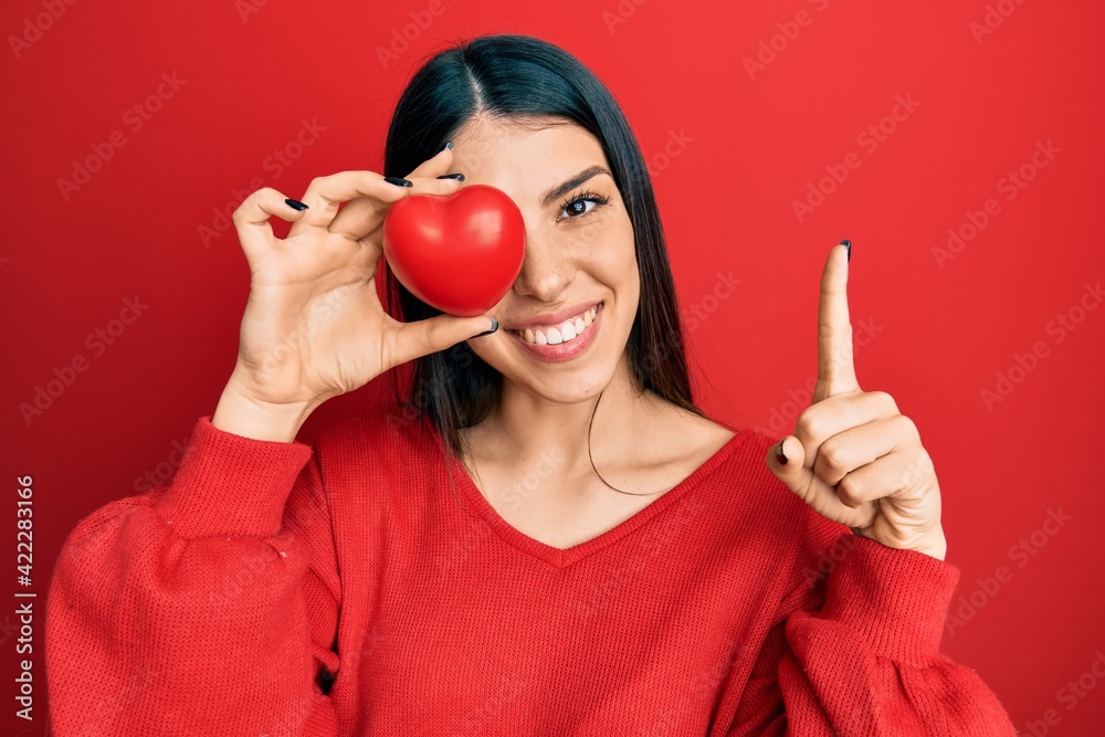 Young hispanic woman holding heart over eye smiling with an idea or question pointing finger with happy face, number one Young hispanic woman holding heart over eye smiling with an idea or question pointing finger with happy face, number one