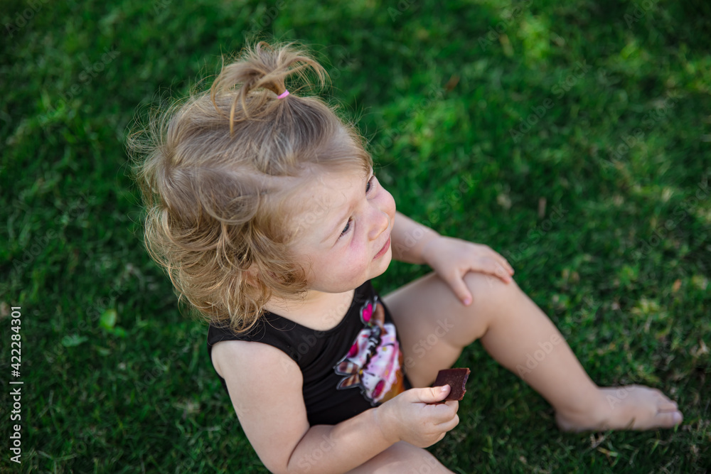 Little funny girl in the summer sits on the grass in the garden with a piece of chocolate in her hand. Little funny girl in the summer sits on the grass in the garden with a piece of chocolate in her hand.