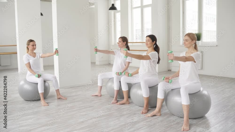 Pregnant females sit on fitballs training with dumbbells, young women in white sportswear engaged in perinatal gymnastics Pregnant females sit on fitballs training with dumbbells, young women in white sportswear engaged in perinatal gymnastics