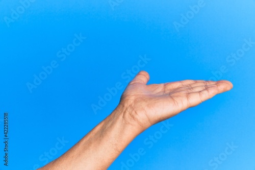 Hand of senior hispanic man over blue isolated background with flat palm presenting product, offer and giving gesture, blank copy space