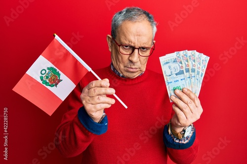 Handsome senior man with grey hair holding peru flag and peruvian sol banknotes skeptic and nervous, frowning upset because of problem. negative person.