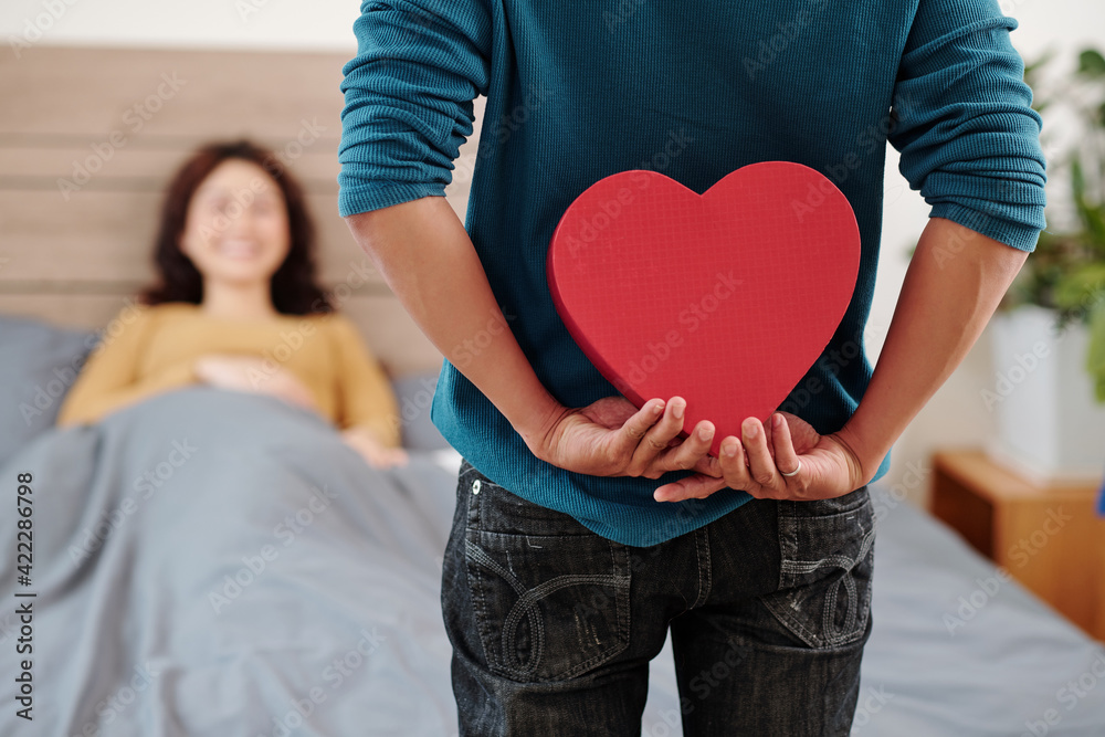 Cropped image of man hiding romantic present behind his back when standing at bed of girlfriend who just woke up Cropped image of man hiding romantic present behind his back when standing at bed of girlfriend who just woke up