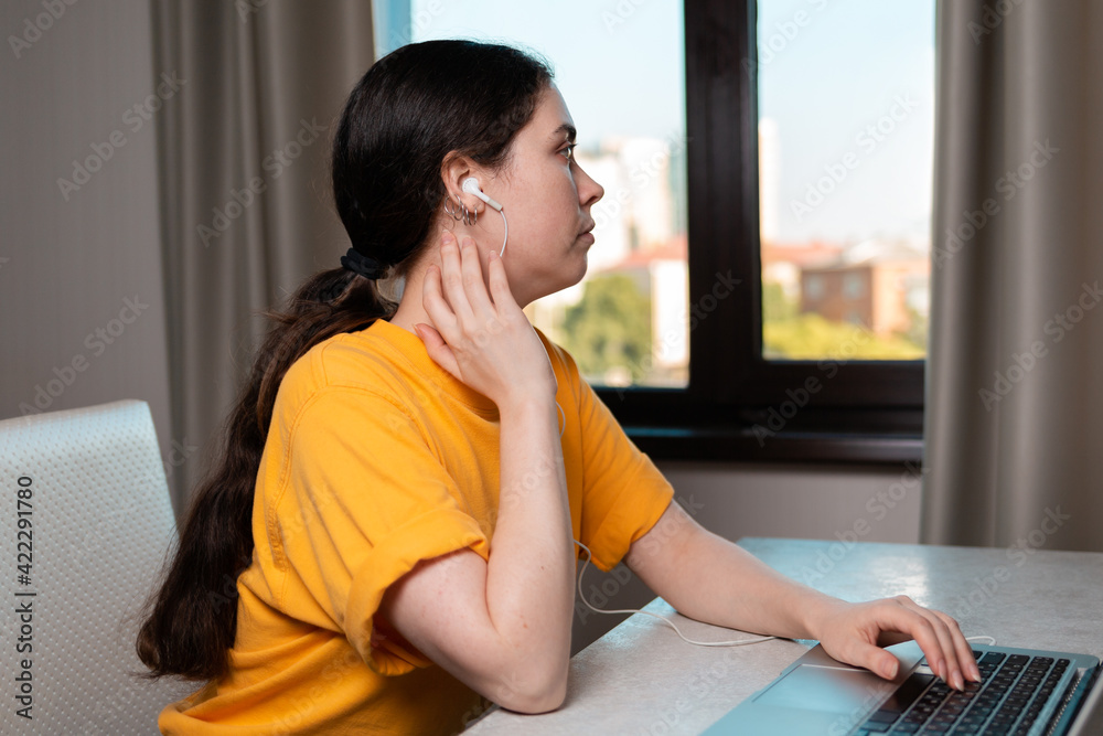 Portrait of a young woman sitting at a desk with headphones on, using a laptop. A window in the background. The concept of online education Portrait of a young woman sitting at a desk with headphones on, using a laptop. A window in the background. The concept of online education