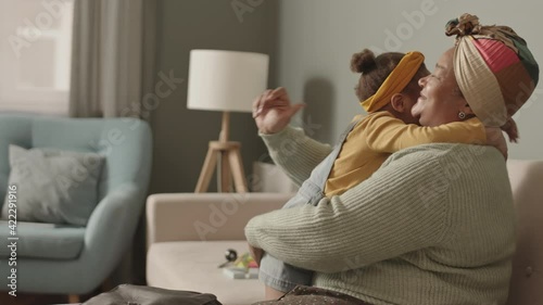 Medium shot of senior African woman sitting on sofa in living room, hugging and kissing on cheek her little adorable granddaughter sitting on her knees photo