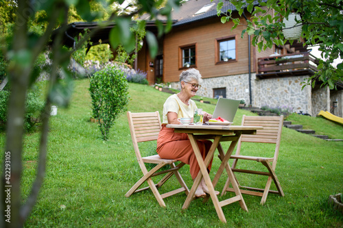 Happy senior woman with laptop working outdoors in garden, home office concept.