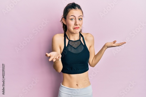 Young brunette woman wearing sportswear clueless and confused expression with arms and hands raised. doubt concept. © Krakenimages.com