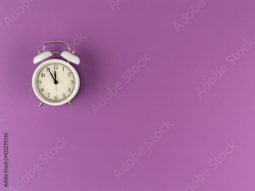 The white alarm clock shows five minutes to twelve on a purple paper background with a copy space. Flat lay. Top view. Place for text. Time: 11.55 am or 23.55 pm
