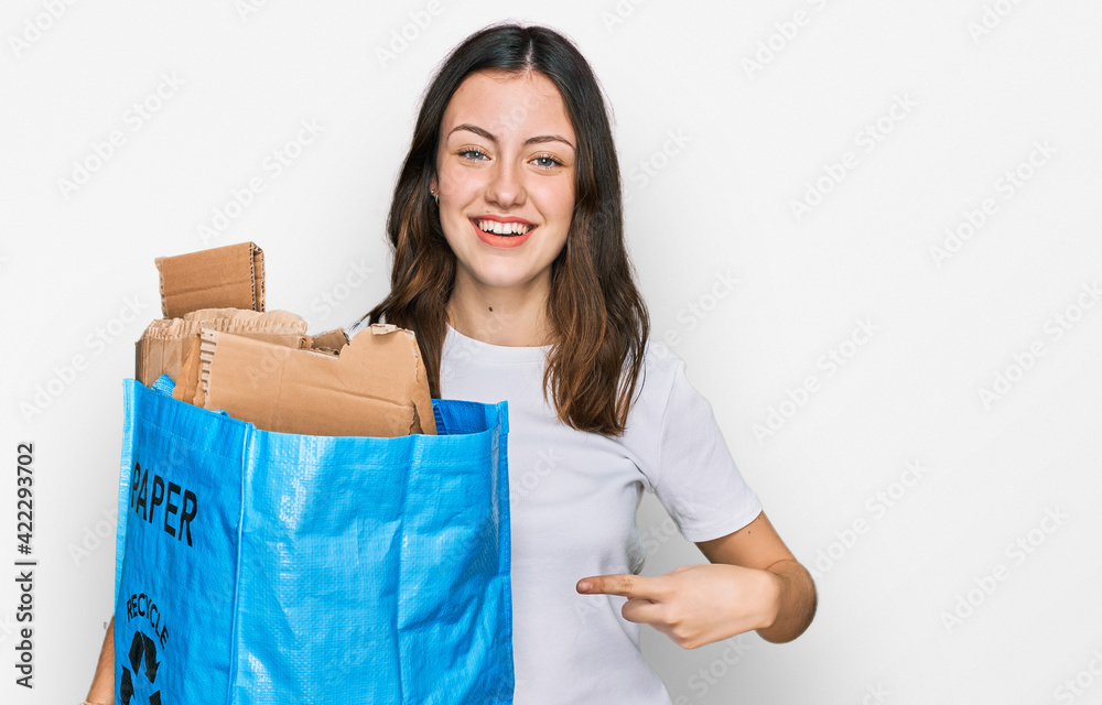 Young beautiful woman holding recycling wastebasket with paper and cardboard smiling happy pointing with hand and finger Young beautiful woman holding recycling wastebasket with paper and cardboard smiling happy pointing with hand and finger