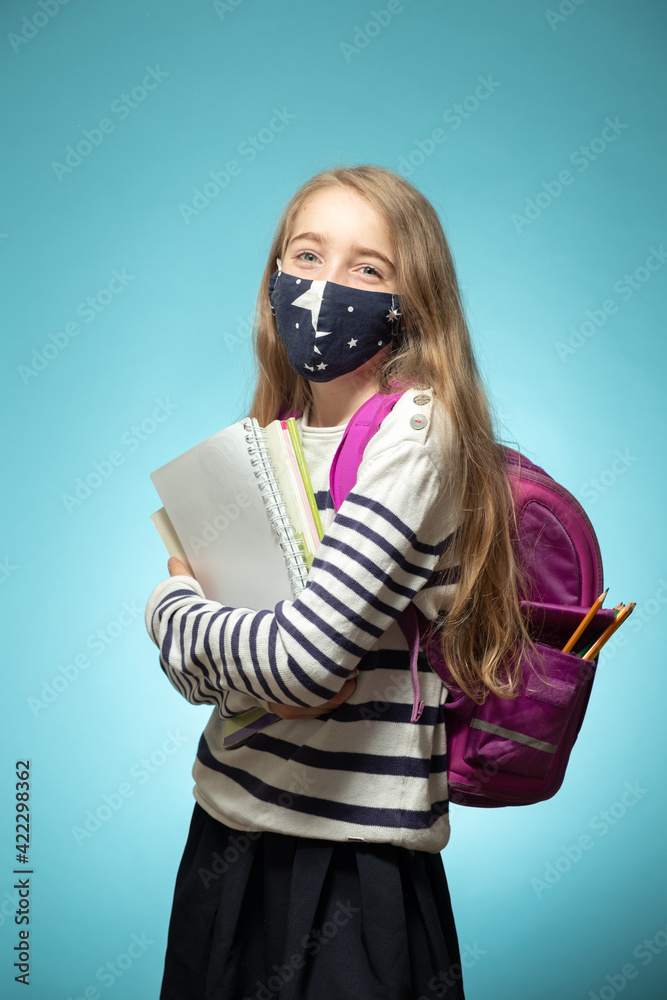 Portrait of a schoolgirl in a protective mask with a backpack on a blue background. The girl is dressed in a school uniform and holds notebooks in her hands. Studio shot. Portrait of a schoolgirl in a protective mask with a backpack on a blue background. The girl is dressed in a school uniform and holds notebooks in her hands. Studio shot.