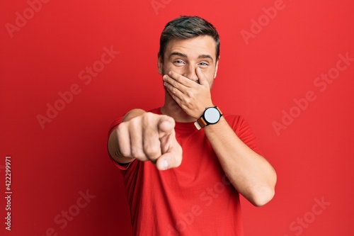 Handsome caucasian man wearing casual red tshirt laughing at you, pointing finger to the camera with hand over mouth, shame expression
