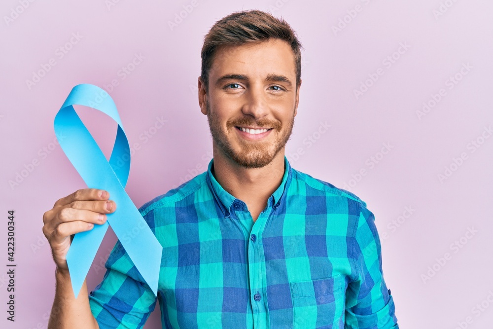 Handsome caucasian man holding blue ribbon looking positive and happy standing and smiling with a confident smile showing teeth Handsome caucasian man holding blue ribbon looking positive and happy standing and smiling with a confident smile showing teeth