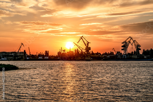 Kherson port at sunset (Ukraine). View from the Dnieper River to the coastline with harbor cranes and commercial ships illuminated by orange sunbeams