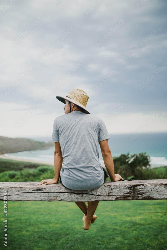 fotografía vertical de un hombre joven con sombrero sentado sobre una viga de madera de un mirador con una playa de fondo, mirando hacia el costado izquierdo - obrazy, fototapety, plakaty 