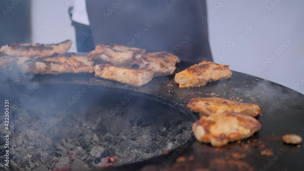 Slow motion: chef holding tongs and grilling fresh juicy meat steaks on round brazier with hot flame at summer local food market - close up view. Outdoor cooking, gastronomy, street food concept Slow motion: chef holding tongs and grilling fresh juicy meat steaks on round brazier with hot flame at summer local food market - close up view. Outdoor cooking, gastronomy, street food concept