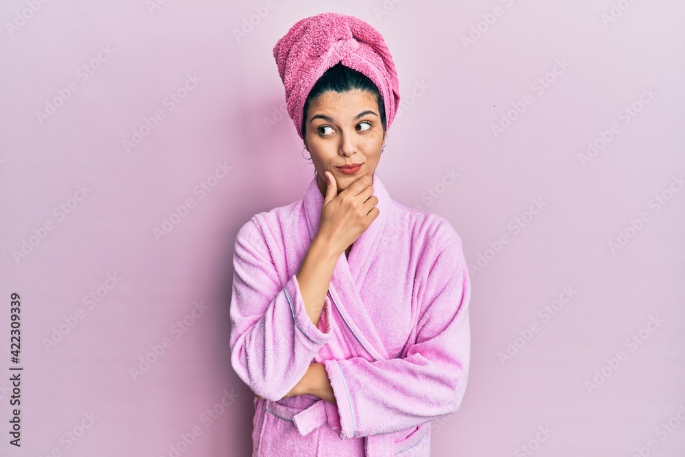 Young hispanic woman wearing shower towel cap and bathrobe with hand on chin thinking about question, pensive expression. smiling with thoughtful face. doubt concept. Young hispanic woman wearing shower towel cap and bathrobe with hand on chin thinking about question, pensive expression. smiling with thoughtful face. doubt concept.