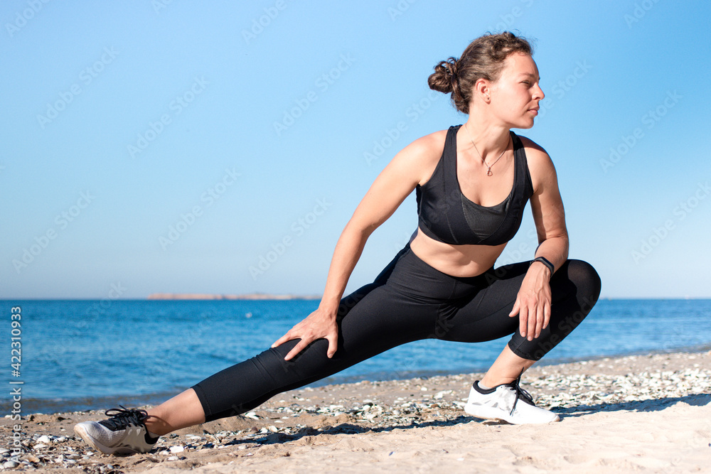 Young brunette athletic woman stretching legs before jogging on the beach in summer morning. Doing lunges exercise outdoors at sea shore. Prepare muscles and ligaments before training workout Young brunette athletic woman stretching legs before jogging on the beach in summer morning. Doing lunges exercise outdoors at sea shore. Prepare muscles and ligaments before training workout
